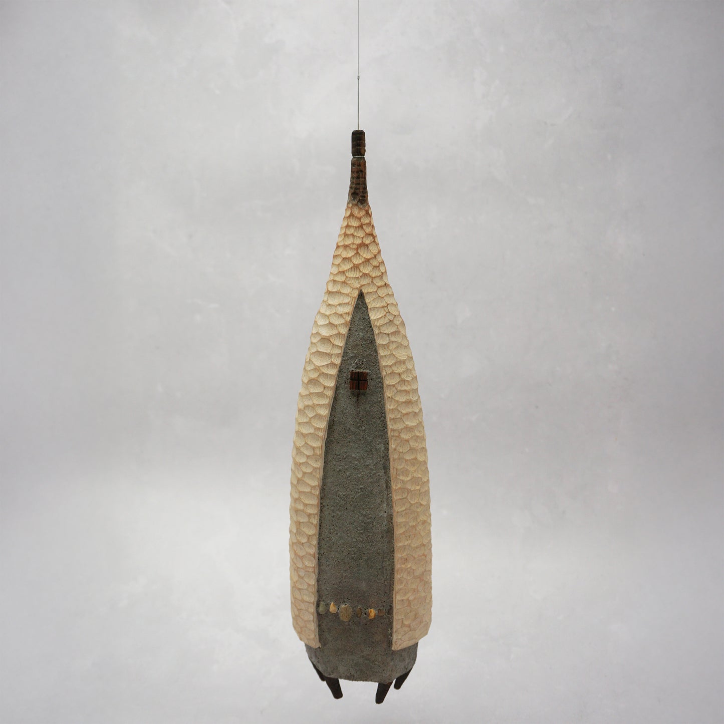 A floating wooden house featuring a hand carved pattern on the sides and back, and a grey mud like colour on the front. The house has four feet and a carved wooden window at the front. The house is hanging from grey thread.