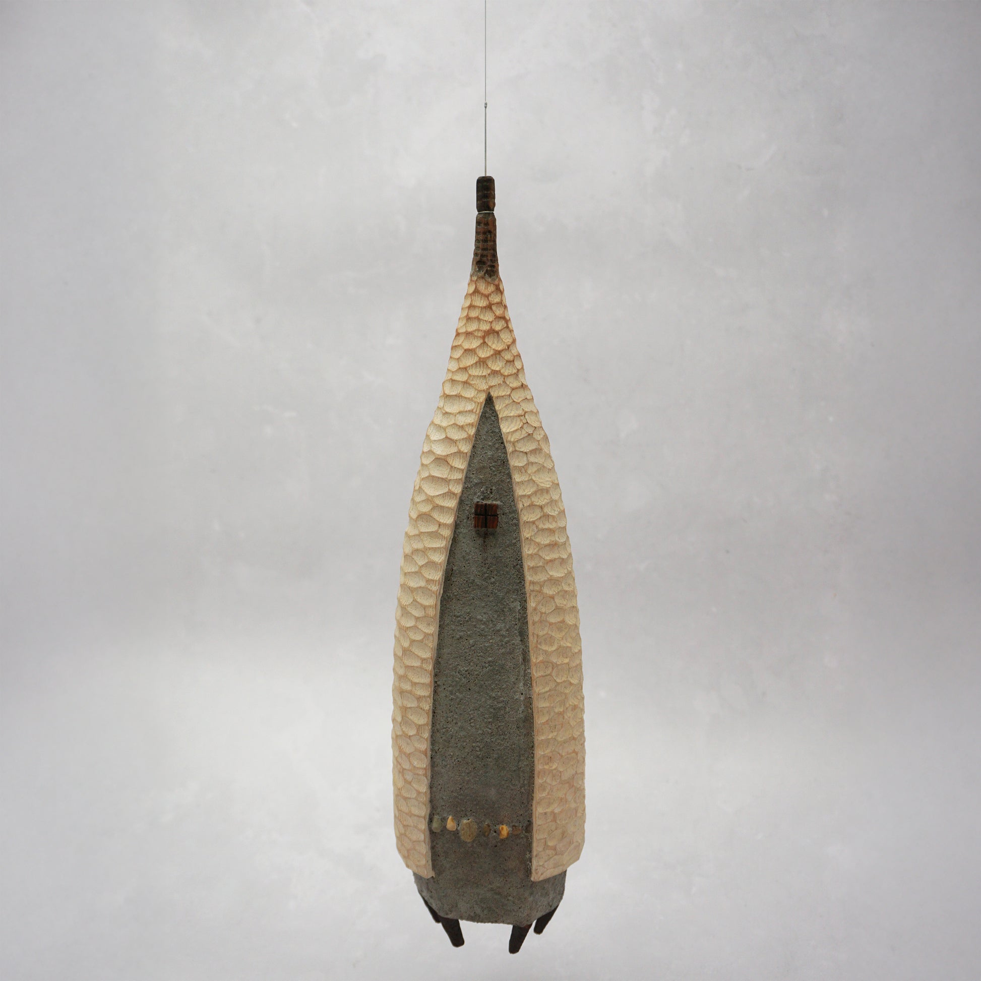 A floating wooden house featuring a hand carved pattern on the sides and back, and a grey mud like colour on the front. The house has four feet and a carved wooden window at the front. The house is hanging from grey thread.