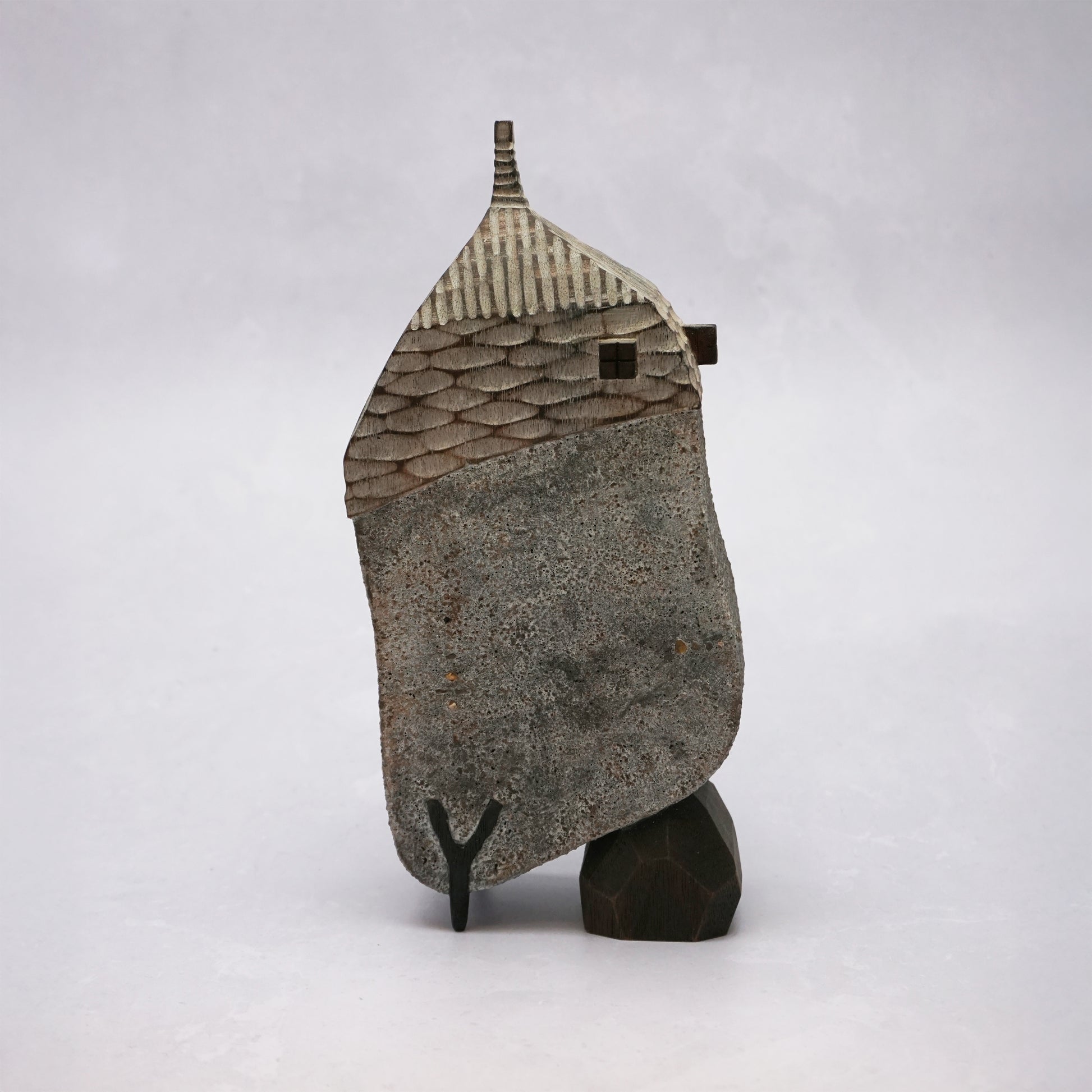 A grey hand carved wooden house with two small legs and a boulder holding it up. The roof is textured and had a small chimney and the house has two small wooden windows.