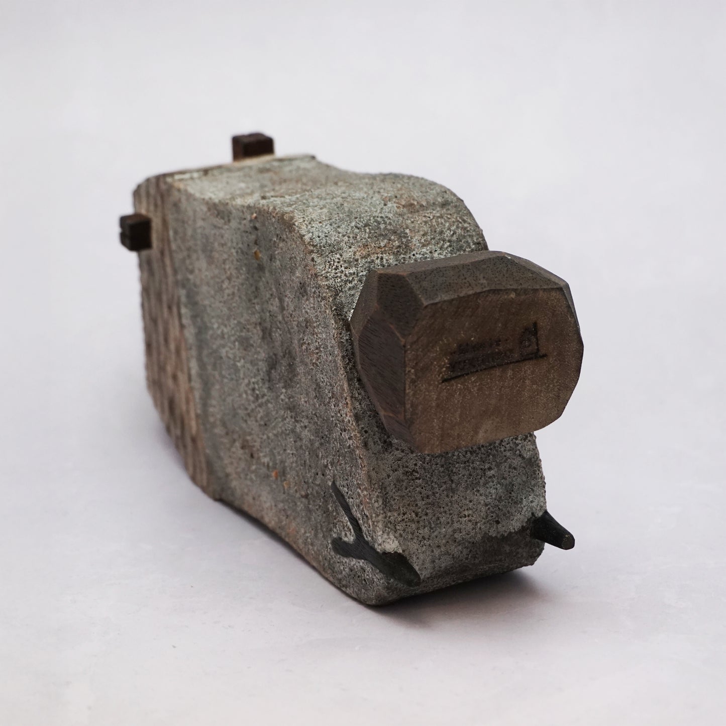 A grey hand carved wooden house with two small legs and a boulder holding it up. The roof is textured and had a small chimney and the house has two small wooden windows.