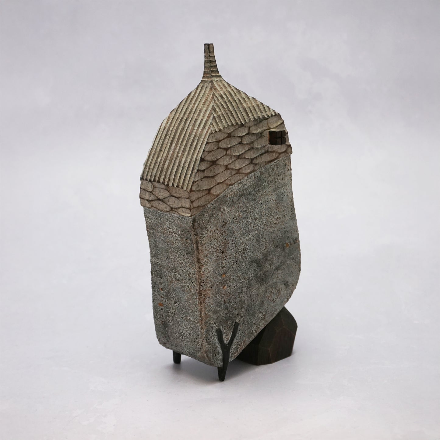 A grey hand carved wooden house with two small legs and a boulder holding it up. The roof is textured and had a small chimney and the house has two small wooden windows.