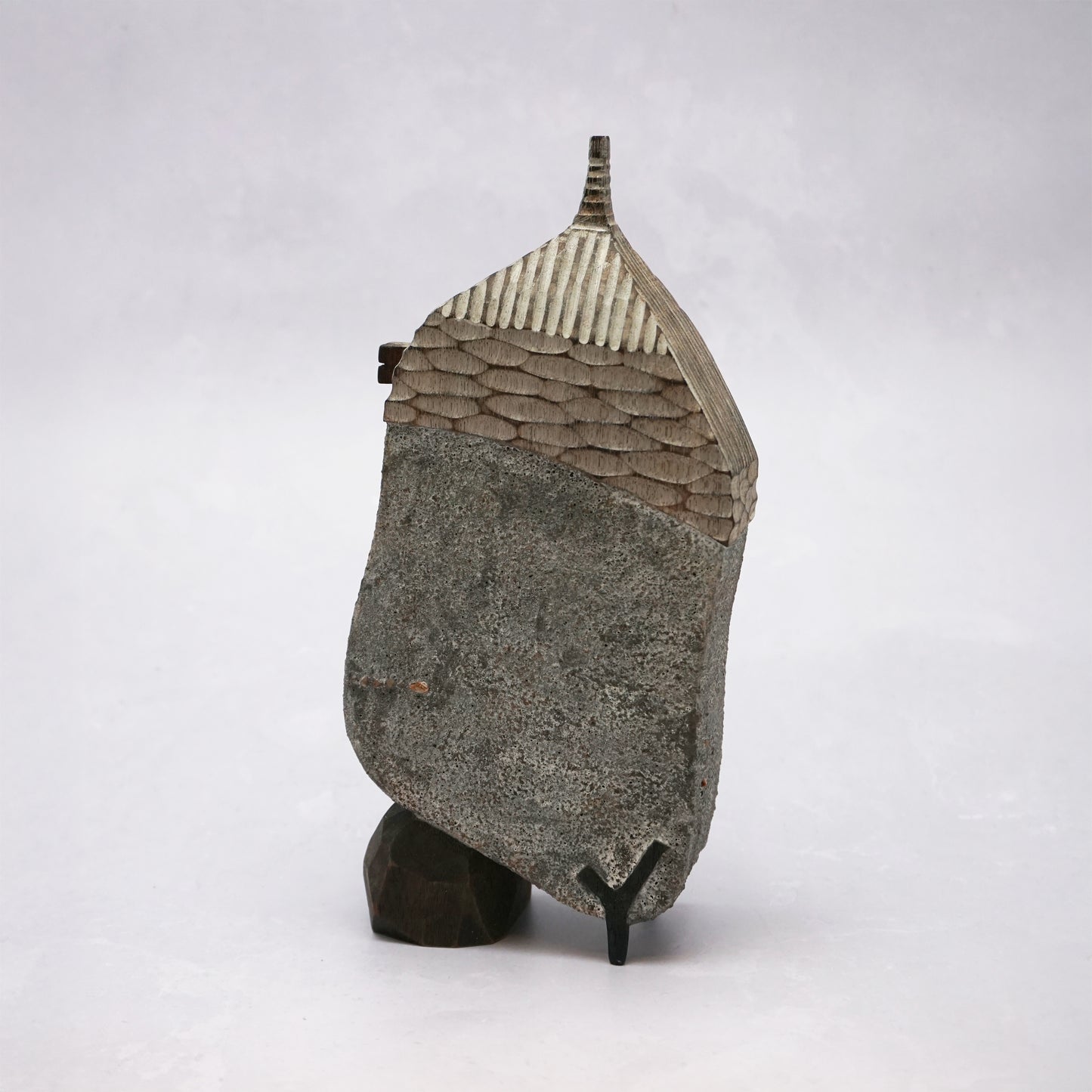 A grey hand carved wooden house with two small legs and a boulder holding it up. The roof is textured and had a small chimney and the house has two small wooden windows.