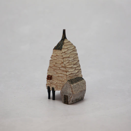 A small hand carved wooden house with a carved textured body, a small chimney and legs at the bottom.