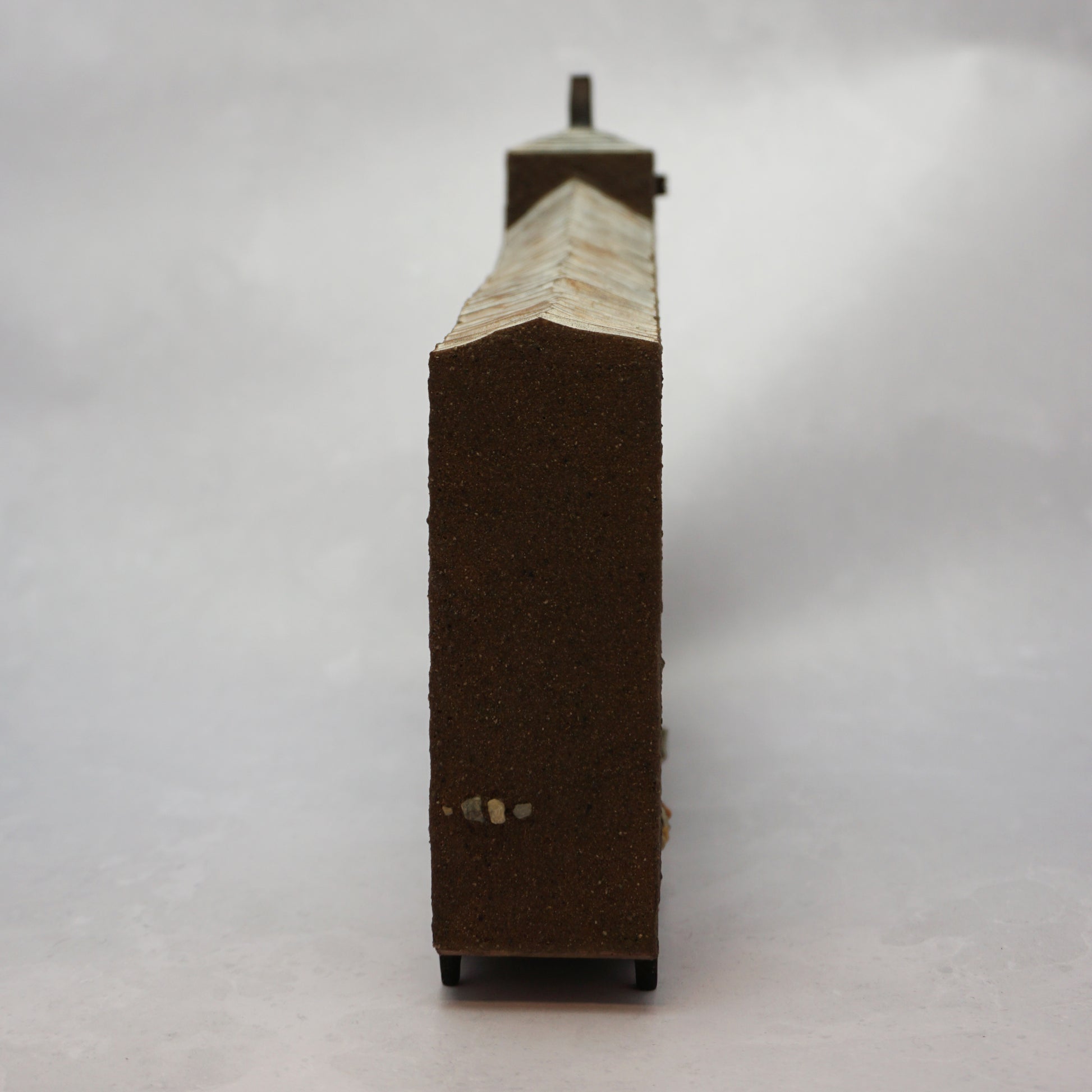 The end of a thin, wide brown wooden house with windows and a carved, textured roof with a small chimney.