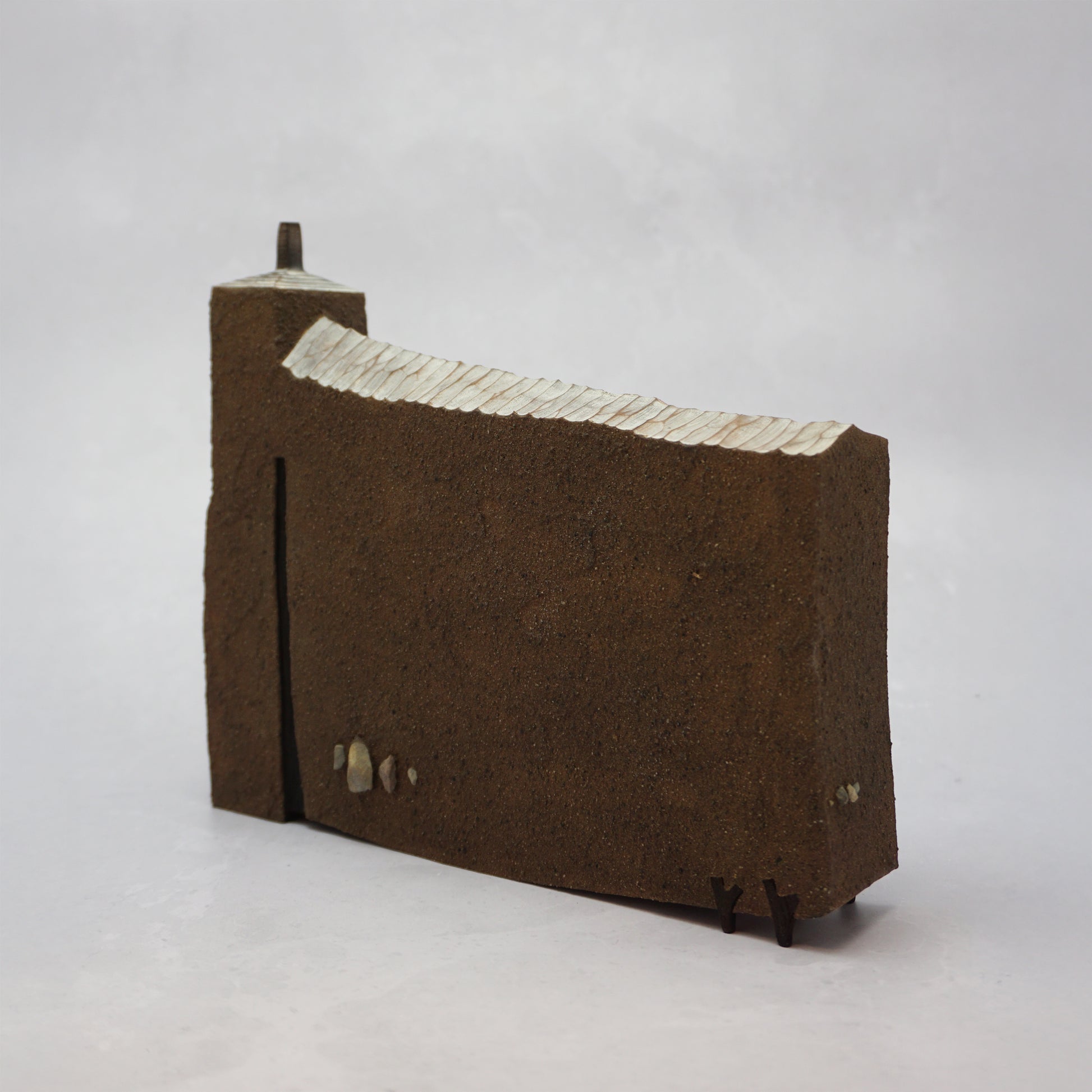 The back of a thin, wide brown wooden house with windows and a carved, textured roof with a small chimney.