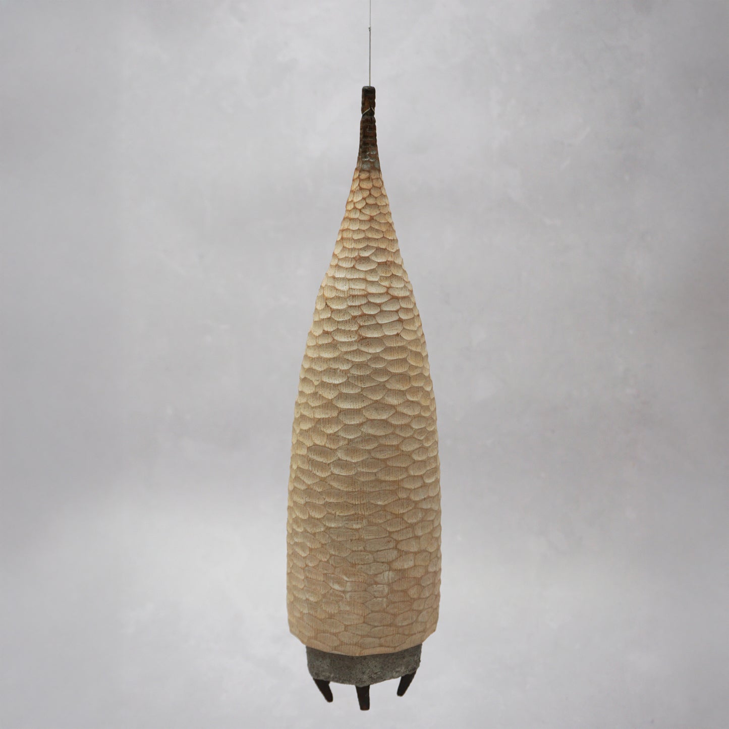 A floating wooden house featuring a hand carved pattern on the sides and back, and a grey mud like colour on the front. The house has four feet and a carved wooden window at the front. The house is hanging from grey thread.