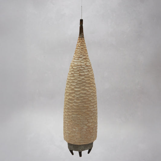 A floating wooden house featuring a hand carved pattern on the sides and back, and a grey mud like colour on the front. The house has four feet and a carved wooden window at the front. The house is hanging from grey thread.