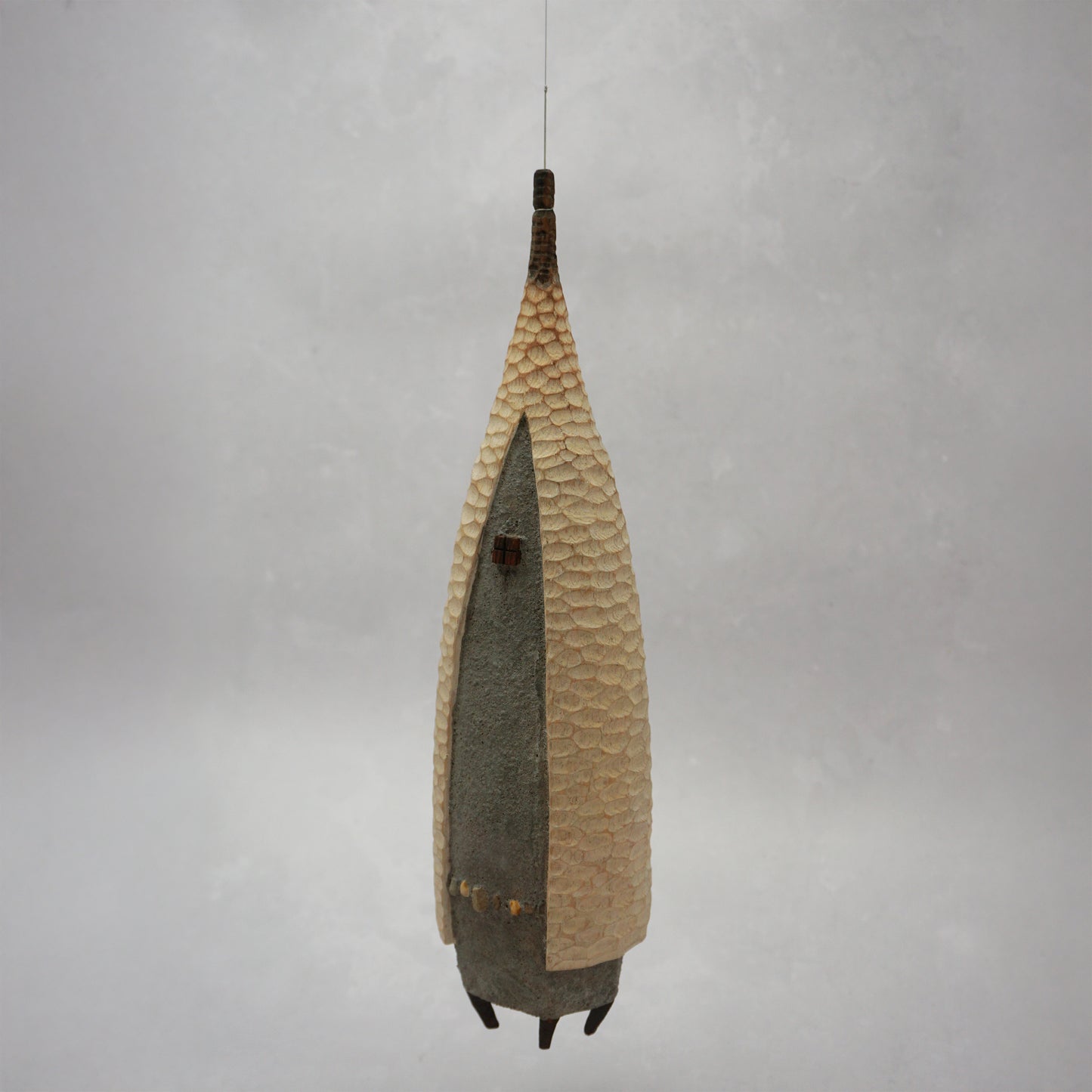 A floating wooden house featuring a hand carved pattern on the sides and back, and a grey mud like colour on the front. The house has four feet and a carved wooden window at the front. The house is hanging from grey thread.