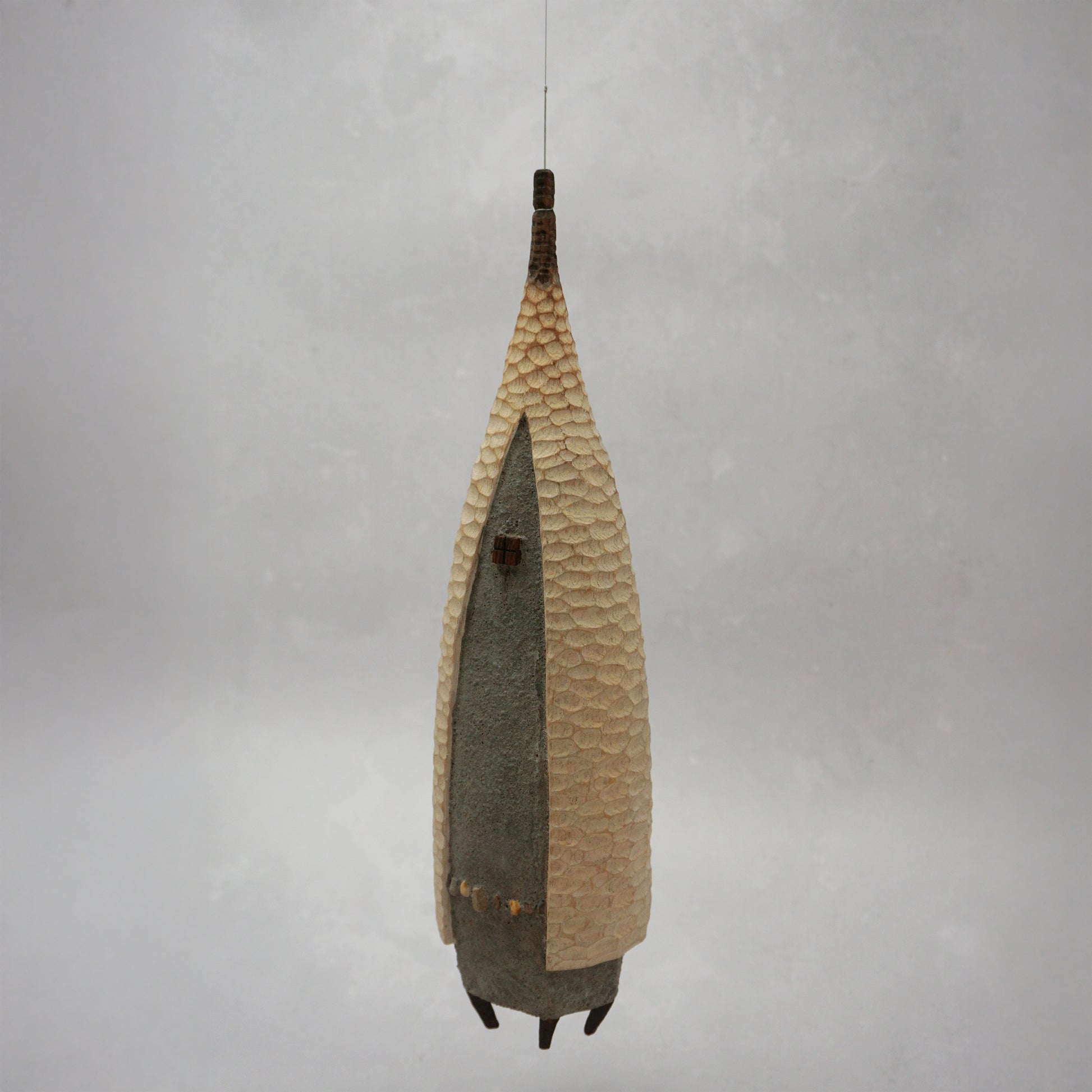 A floating wooden house featuring a hand carved pattern on the sides and back, and a grey mud like colour on the front. The house has four feet and a carved wooden window at the front. The house is hanging from grey thread.