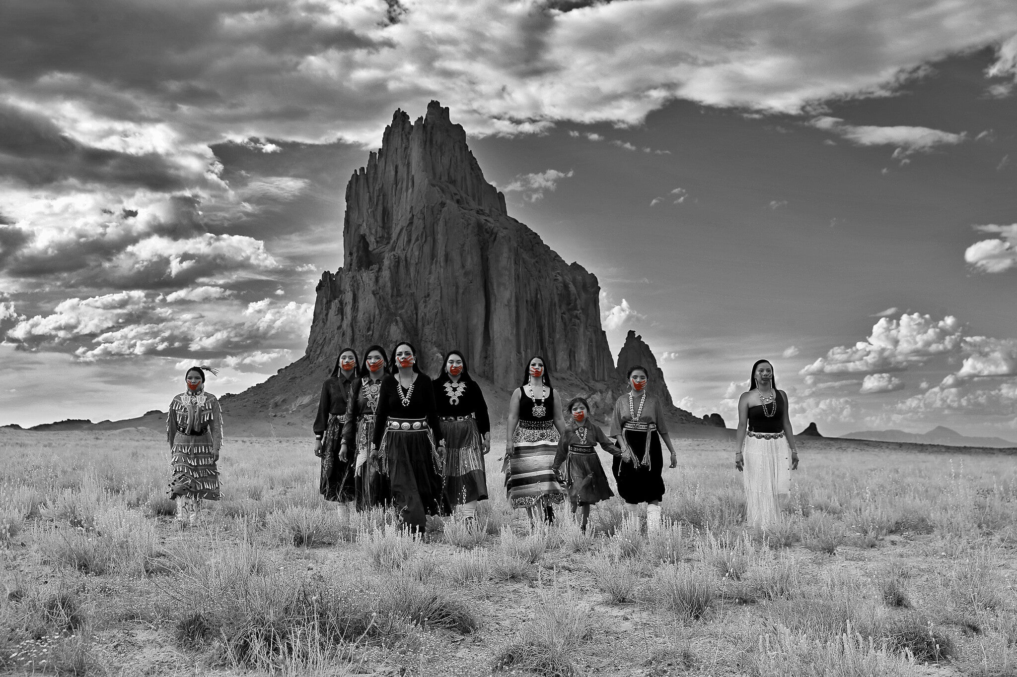 A group of eight women dressed in traditional attire walks towards the camera in a vast, open landscape. The background features a prominent rock formation under a dramatic sky, with clouds casting shadows over the scene. The image is in black and white, highlighting the women's vibrant red lips.