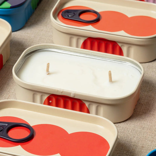 An open beige tin with red spots with a double wick candle inside surrounded by other similar tins.