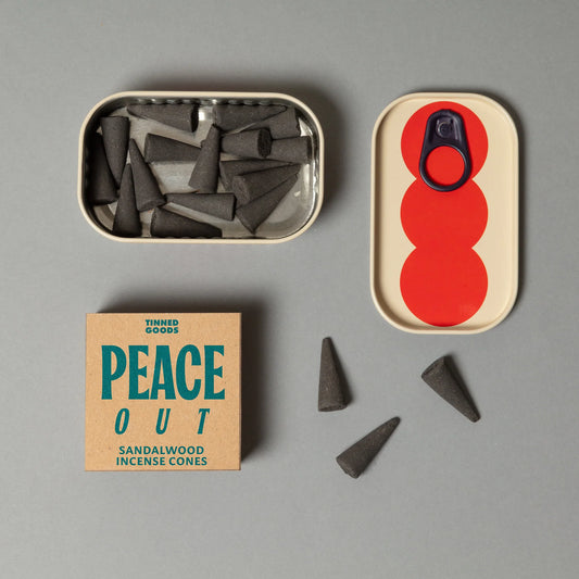 A flat lay image of an pen tin grey of incense cones, a beige tin lid featuring red circles, three loose incense cones and a brown cardboard square featuring the words 'Peace Out Sandalwood Incense Cones'.