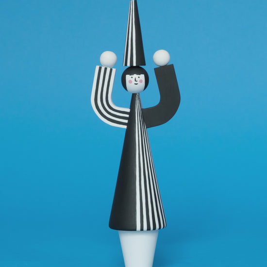 A spinning video of a wooden sculpture with a black and white stripey body with a matching headdress featuring a blue pompom on the top with white legs on a plain blue background.
