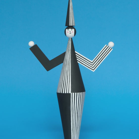 A spinning video of a wooden sculpture with a black and white stripey body with a matching headdress featuring a blue pompom on the top with white and black stripey legs on a plain blue background.