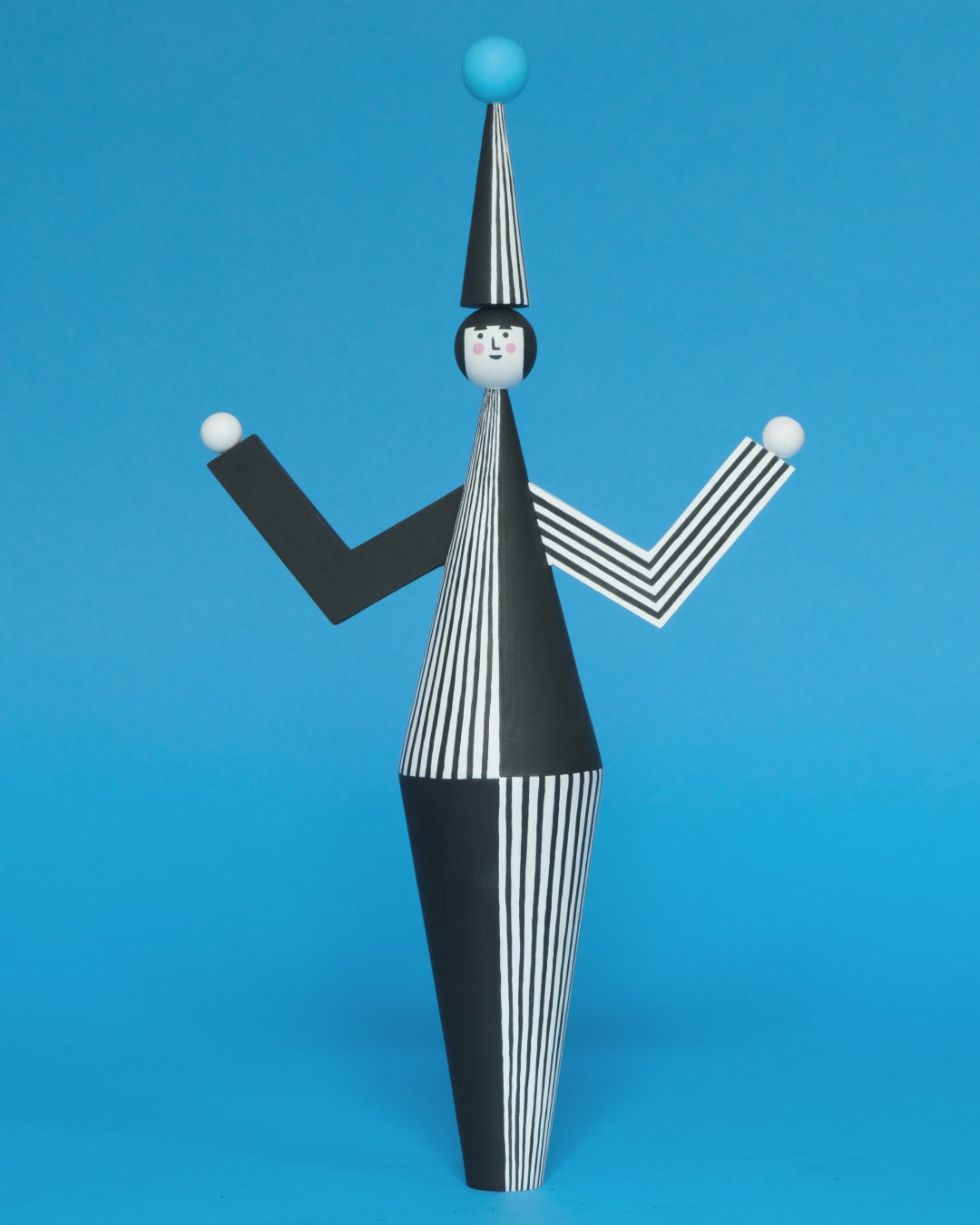 A spinning video of a wooden sculpture with a black and white stripey body with a matching headdress featuring a blue pompom on the top with white and black stripey legs on a plain blue background.