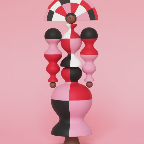 A spinning video of a colourful sculpture with a white, red, black and pink stripey curved body with a matching headdress on a plain pink background.