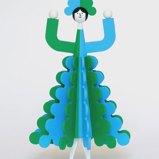 A spinning video of a colourful wooden sculpture with a green and blue stripey 3D skirt and a matching headdress with white legs on a plain white background.