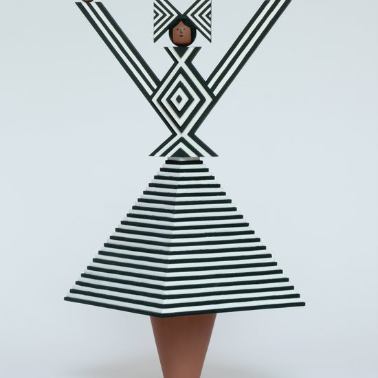 A spinning video of a monochrome wooden sculpture with a black and white stripey 3D top and skirt and a matching headdress with brown legs on a plain white background.