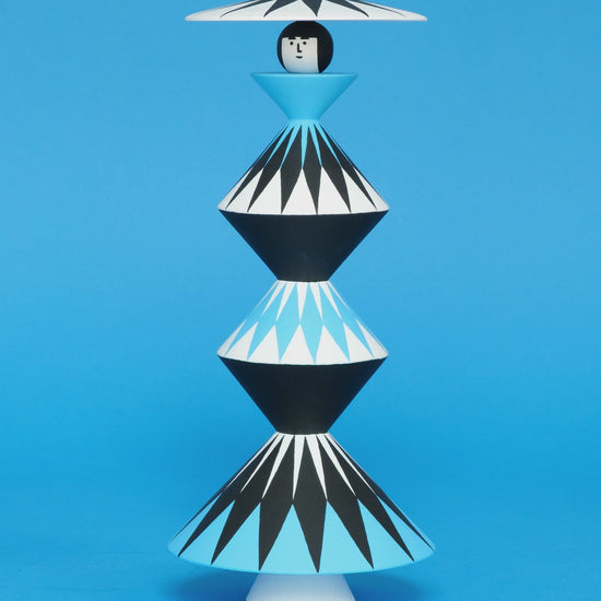 A spinning video of a colourful wooden sculpture with a black, white and blue diamond patterned triangular stacked body with white legs and a matching headdress on a plain blue background.