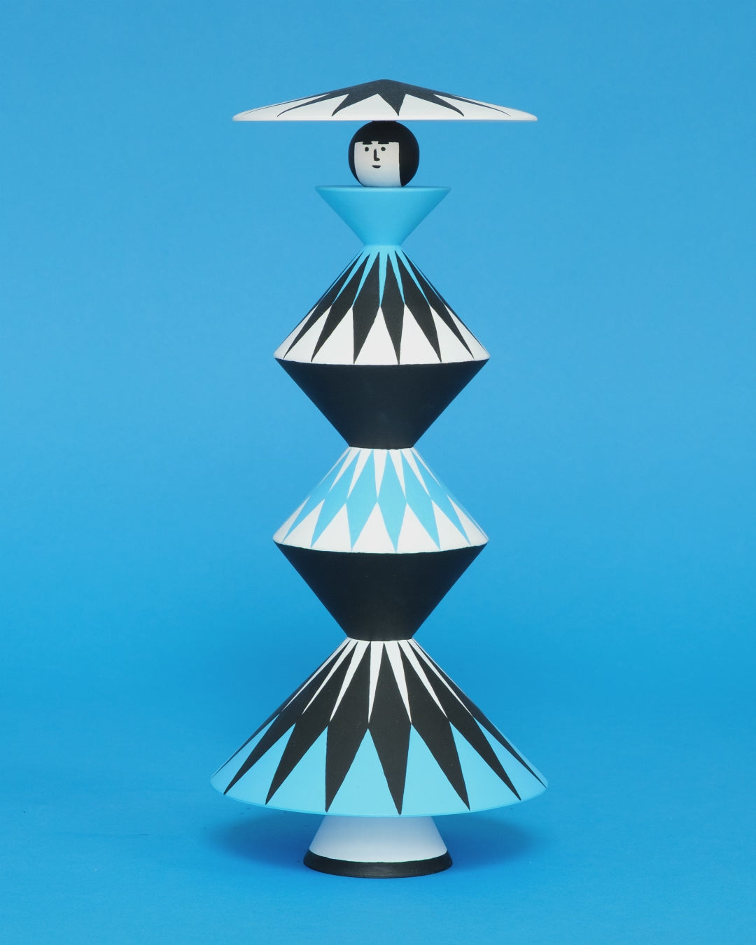 A spinning video of a colourful wooden sculpture with a black, white and blue diamond patterned triangular stacked body with white legs and a matching headdress on a plain blue background.