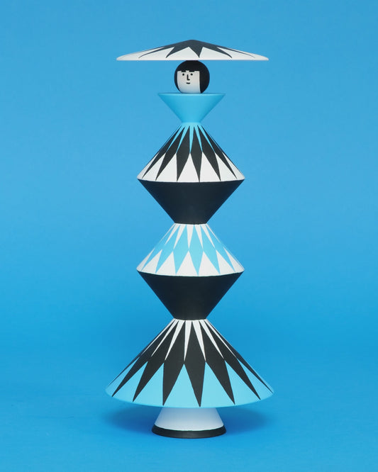 A spinning video of a colourful wooden sculpture with a black, white and blue diamond patterned triangular stacked body with white legs and a matching headdress on a plain blue background.