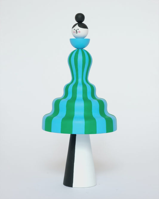 A spinning video of a colourful sculpture of a figure made from a green and blue stripey body with white and black legs on a plain white background.