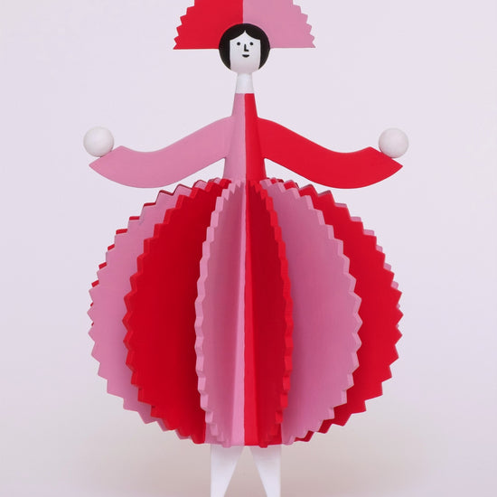 A spinning video of a colourful wooden sculpture with a pink and red stripey 3d round dress and a matching headdress and white legs on a plain white background.