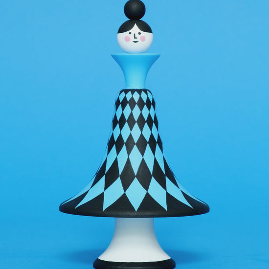A spinning video of a colourful wooden sculpture with a black and blue diamond patterned curved body with white legs on a plain blue background.