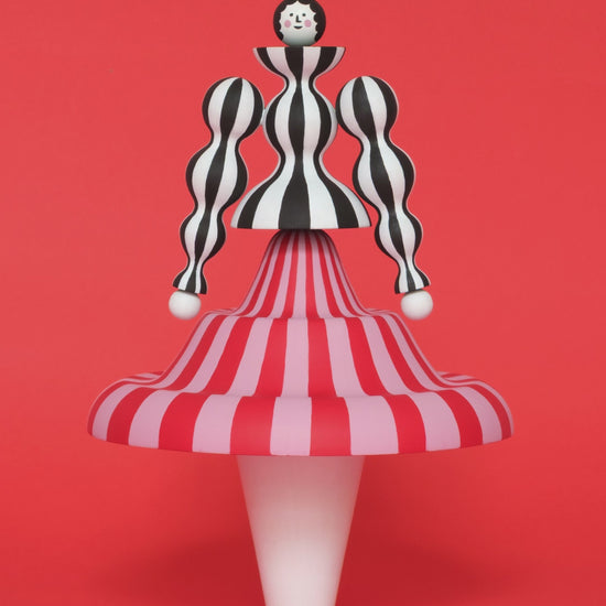 A spinning video of a colourful sculpture of a figure made from a black and white stripey body with a red and pink stripey skirt with white legs on a plain orange background.