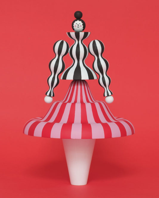 A spinning video of a colourful sculpture of a figure made from a black and white stripey body with a red and pink stripey skirt with white legs on a plain orange background.