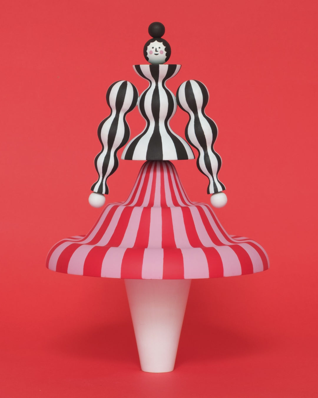 A spinning video of a colourful sculpture of a figure made from a black and white stripey body with a red and pink stripey skirt with white legs on a plain orange background.