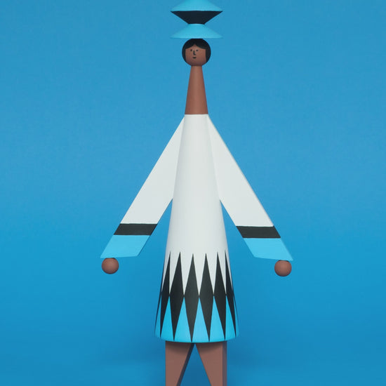 A spinning video of a colourful wooden sculpture with a black, white and blue diamond patterned dress with brown legs and a matching blue and black headdress on a plain blue background.