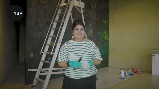 Portrait of Kéyah Yinílínih (The and Remembers) painting a large, textured artwork. A paint brush and palette are visible in the foreground.