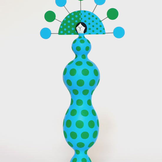 A spinning video of a colourful sculpture with a blue and green spotty body with a matching headdress on a plain white background.