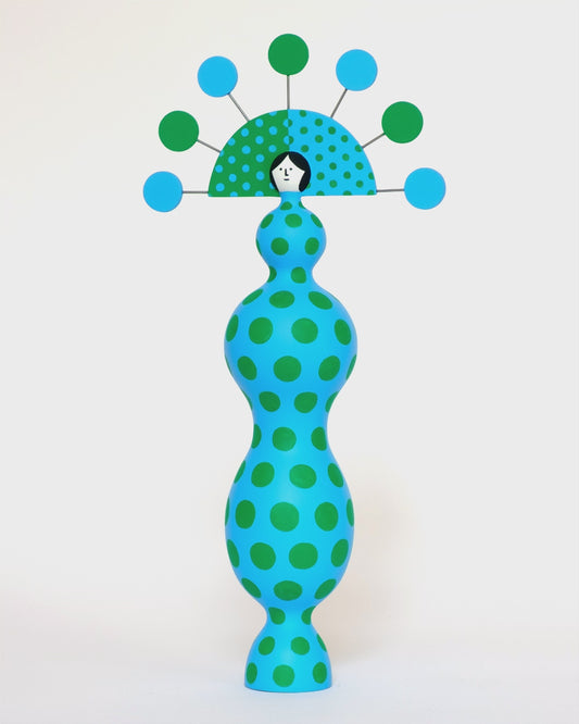 A spinning video of a colourful sculpture with a blue and green spotty body with a matching headdress on a plain white background.