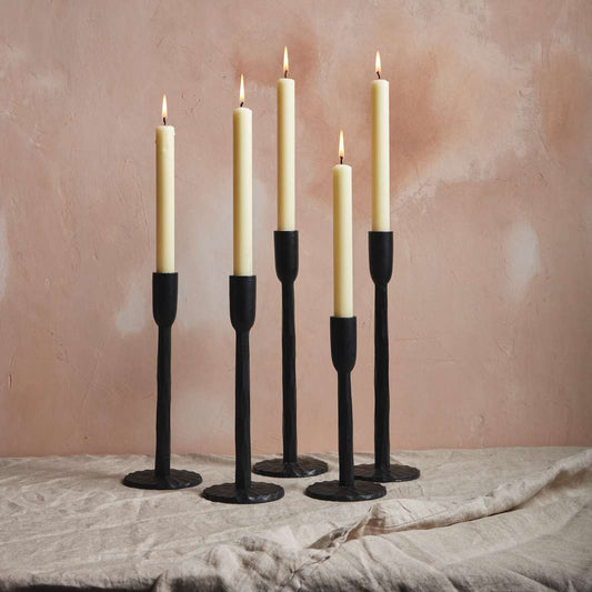 Five tall, slender candles with cream-coloured wax are lit and placed in black candle holders. They stand on a light, textured fabric against a soft, peach-toned wall, creating a warm and inviting atmosphere.