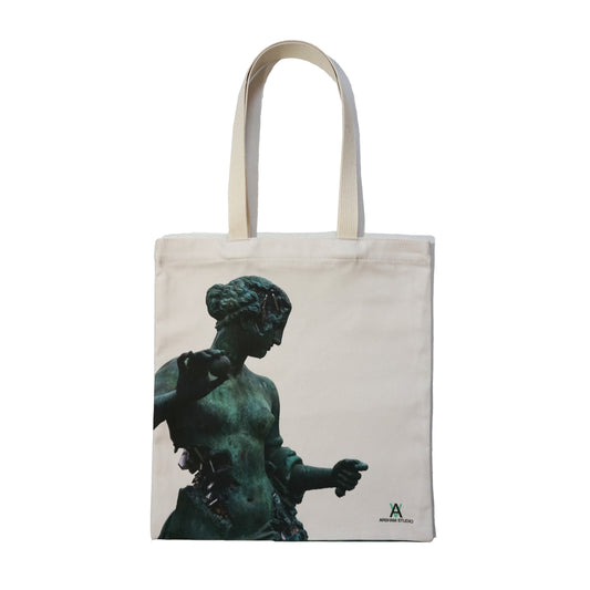 A canvas tote bag featuring a silhouette of a classical statue, depicted in a greenish hue. The statue appears to be a female figure, elegantly posed, with intricate details visible. The bag has two sturdy handles and a logo at the bottom.