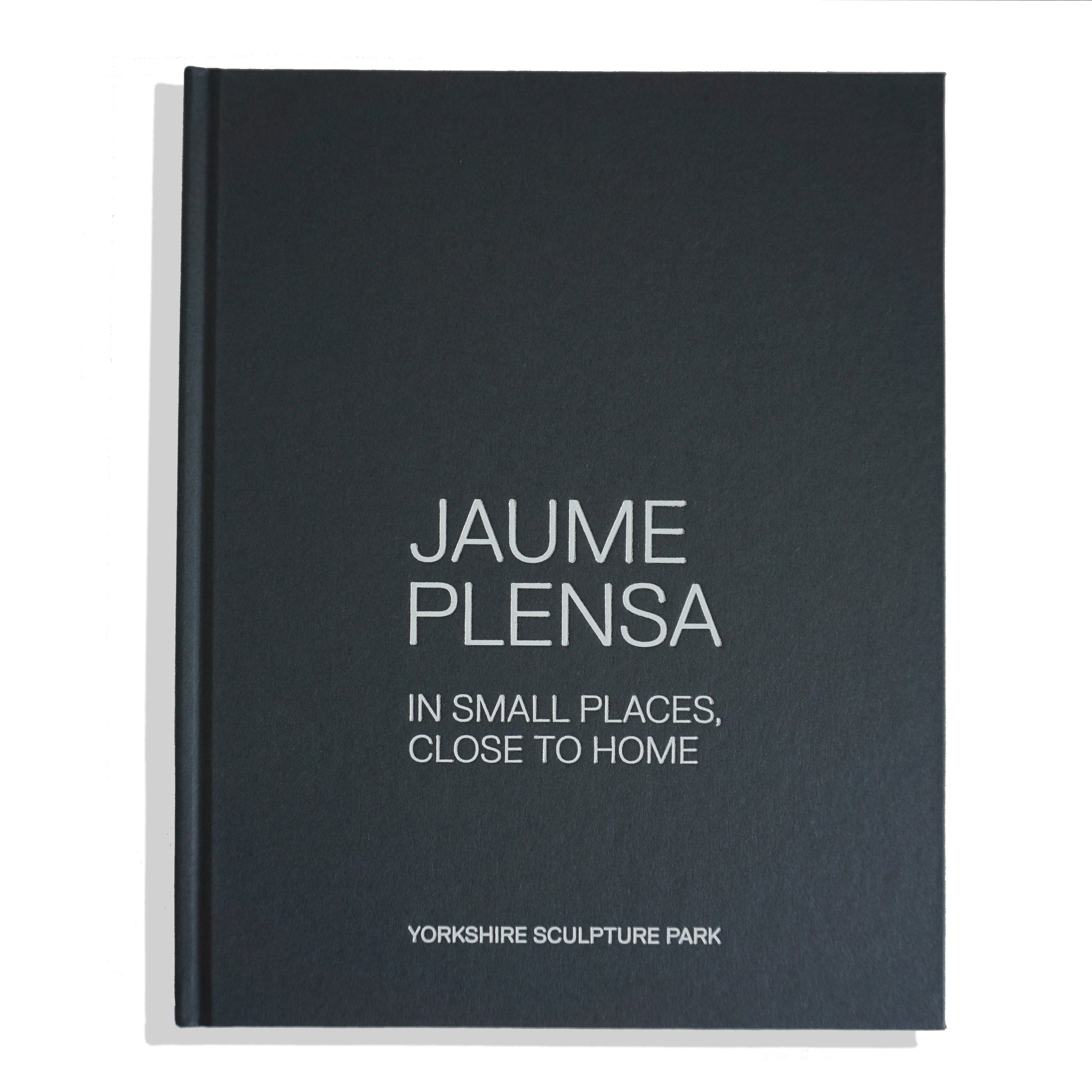 A closed book titled "Jaume Plensa: In Small Places, Close to Home" with a dark green, blue cover and white text. The book is associated with the Yorkshire Sculpture Park, suggesting a focus on the artist's work and themes.