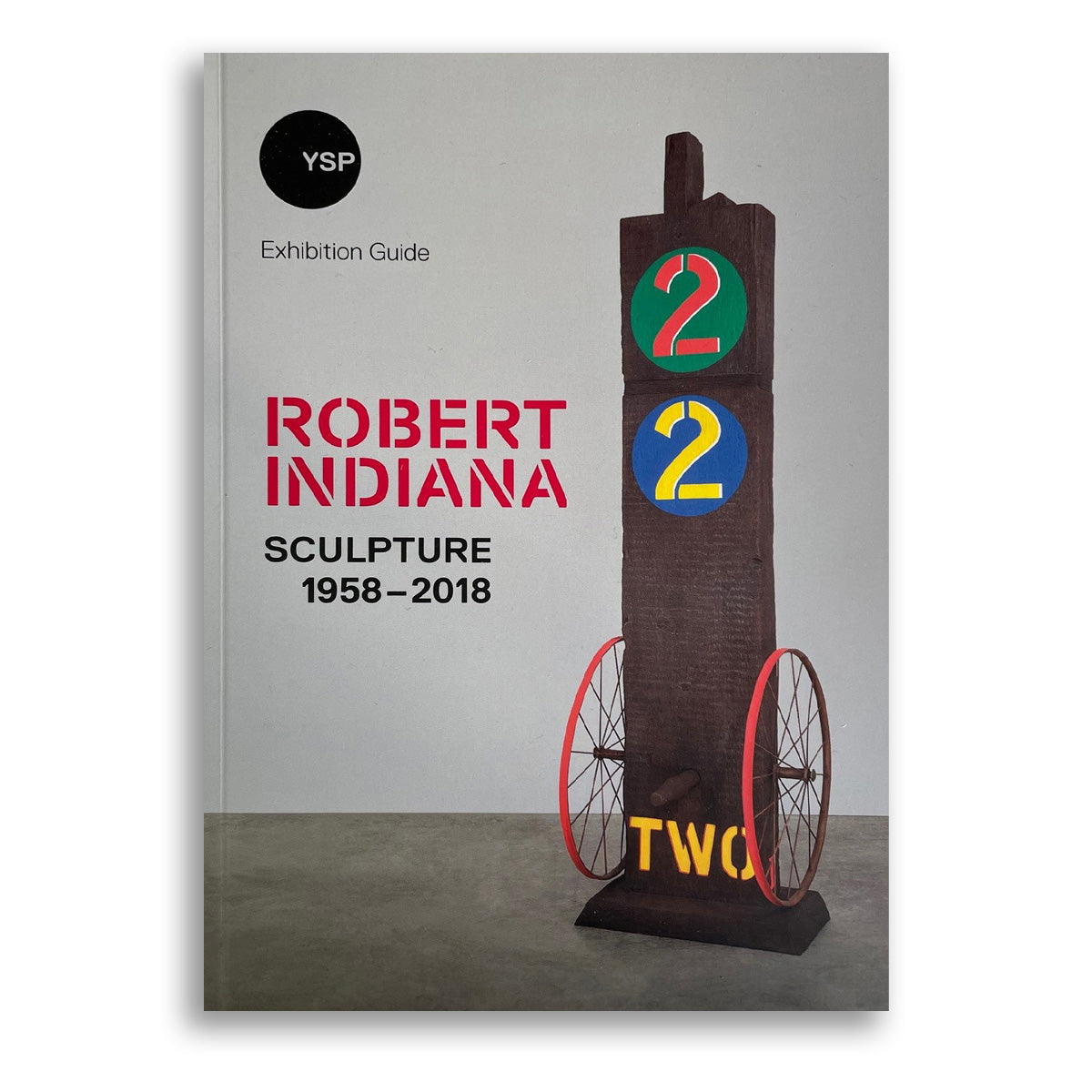 A book cover featuring the title "Robert Indiana: Sculpture 1958–2018" in bold red text. The cover displays a wooden sculpture with the numbers "2" and "2" in green and blue, alongside the word "TWO" in yellow, set against a grey background.